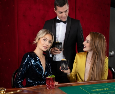 Two women are seated at a gaming table with a roulette and a RIEDEL Laudon Red glass on it while one of them holds a RIEDEL Laudon Black glass. Behind them is a man in front of a red background who is also holding a RIEDEL Laudon Black glass. 