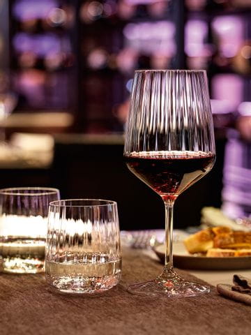 The SPIEGELAU Lifestyle series on a restaurant table in a cosy evening atmosphere. Purple light shines through the smooth wave structure of the filled white wine glass, red wine glass, water tumblers and carafe.<br/>