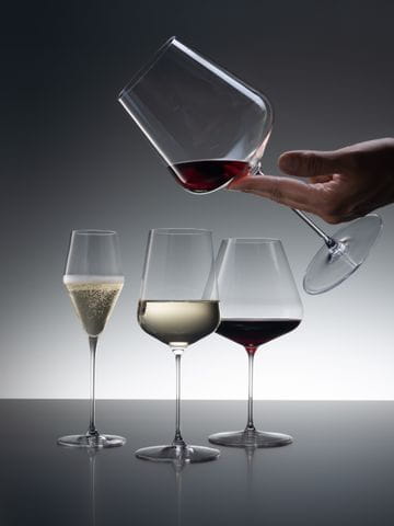 Drei SPIEGELAU Definition Gläser, von denen das Champagnerglas mit Sekt, das Universalglas mit Weißwein und das Burgunderglas mit Rotwein gefüllt ist. Von der rechten oberen Ecke kommend hält eine Hand das Bordeaux-Glas, das mit etwas Rotwein gefüllt ist.<br/>
