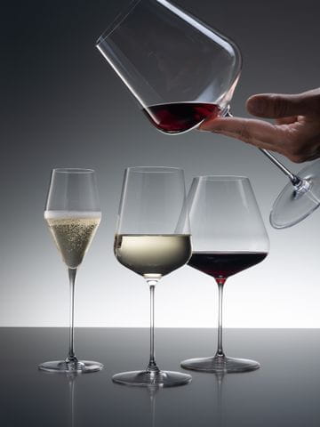 Three SPIEGELAU Definition glasses of which the Champagne glass is filled with sparkling wine, the Universal glass with white wine and the Burgundy glass with red wine. Coming from the top right corner, a hand is holding the Bordeaux glass filled with a little red wine.<br/>