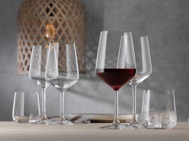 Die Glasserie SPIEGELAU Style auf einem Tisch. Im Vordergrund stehen ein mit Rotwein gefülltes Rotweinglas, ein mit Wasser gefüllter Becher und ein leeres Weißweinglas. Im Hintergrund stehen die gleichen Gläser, allerdings leer, sowie eine korbartige Deckenlampe.<br/>