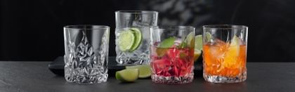 Quattro bicchieri da whisky NACHTMANN Sculpture su un tavolo, tre dei quali riempiti con diverse bevande alcoliche e analcoliche. Tra i bicchieri il tavolo è decorato con fette di lime.<br/>