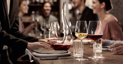 The filled white wine glass and the filled Bordeaux glass SPIEGELAU Willsberger Anniversary in front of the SPIEGELAU Perfect Serve Collection SOF glass and the filled SPIEGELAU Novo decanter. All of them on a dinner table surrounded by people.<br/>
