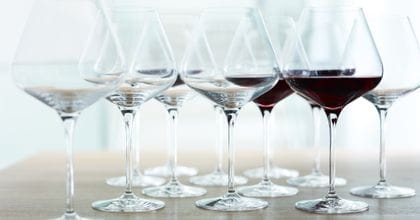 A group of SPIEGELAU Hybrid Burgundy glasses on a wooden table. Some of the glasses are filled with red wine, some are empty.<br/>