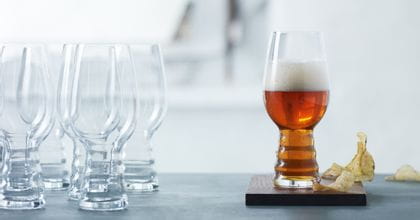 A group of SPIEGELAU Craft Beer Glasses for IPA on a table. One standing on a wooden coaster, filled with IPA beer. Next to this glass are some potato chips.<br/>