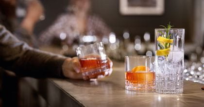 Un bancone da bar con due bicchieri SPIEGELAU Perfect Serve Collection. Il bicchiere SOF è riempito con una bevanda color arancio e una scorza d'arancia, il bicchiere longdrink è riempito con una bevanda da cocktail chiara, cubetti di ghiaccio, rosmarino e una scorza di limone. Sullo sfondo una mano che tiene un altro bicchiere SOF pieno.<br/>