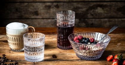 La serie de vasos de cristal NACHTMANN Ethno sobre una mesa de madera decorada con granos de café y fresas. La taza de bebida caliente está llena de capuchino, el vaso de agua mineral, el vaso largo de zumo de bayas rojo oscuro y el cuenco de muesly crujiente.<br/>