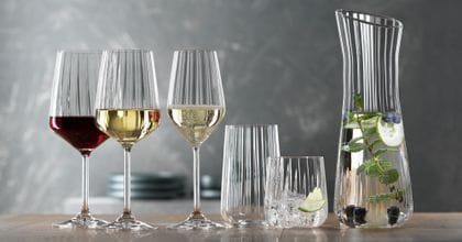 La série SPIEGELAU Lifestyle sur une table. Le verre à vin rouge rempli, le verre à vin blanc rempli, la coupe à champagne remplie, un verre à long drink vide, un gobelet rempli d'eau, de glaçons et d'une tranche de citron vert. À droite, la carafe contenant de l'eau infusée.<br/>
