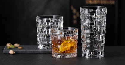 Il NACHTMANN Bossa Nova SOF riempito di whisky accanto al bicchiere longdrink Bossa Nova vuoto su una credenza in pietra nera. Dietro di loro il tumbler Bossa Nova vuoto.<br/>
