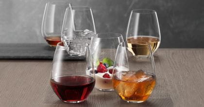 Sechs SPIEGELAU Authentis Casual Becher auf einem Holztisch, jeder von ihnen unterschiedlich gefüllt. Mit Rotwein, mit einem Softdrink auf Eis, mit einem cremigen Dessert, mit Weißwein, mit Wasser und Eiswürfeln und mit einem braunen Likör.<br/>
