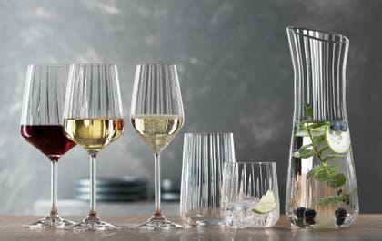 Die SPIEGELAU Lifestyle-Serie auf einem Tisch. Das gefüllte Rotweinglas, das gefüllte Weißweinglas, das gefüllte Champagnerglas, ein leeres Longdrinkglas, ein Becher gefüllt mit Wasser, Eiswürfeln und einer Limettenscheibe. Rechts die Karaffe mit gefülltem Wasser.<br/>