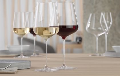 Un verre à bordeaux SPIEGELAU Definition rempli de vin rouge et un verre à vin blanc SPIEGELAU Definition rempli de vin blanc sur une table. À l'arrière-plan, les mêmes verres à vin sont de nouveau remplis.<br/>