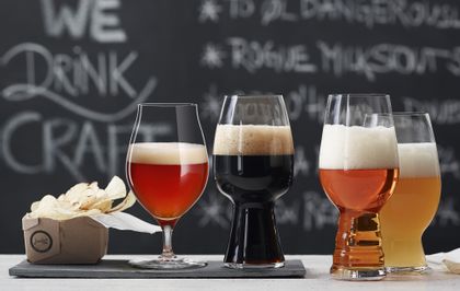 Die mit Bier gefüllten SPIEGELAU Craft Beer Gläser stehen auf einem Tisch vor einer Tafel mit einer Speisekarte. Das Glas für Barrel Aged Bier und das Stout Glas stehen auf einer Schieferplatte, das IPA Glas und das American Wheat Beer Glas daneben auf dem Tisch.<br/>