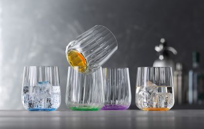 The SPIEGELAU Liefestyle cups in the different colours ocean, leaf, sun and lilac stand in a row. The cup in the colour ocean and sun is filled with water and ice cubes.<br/>