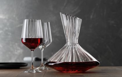 Die mit Rotwein gefüllte SPIEGELAU Lifestyle Karaffe auf einem Tisch. Links von der Karaffe zwei Lifestyle Rotweingläser, eines davon mit Rotwein gefüllt. Am linken Rand des Tisches eine leere schwarze Platte.<br/>