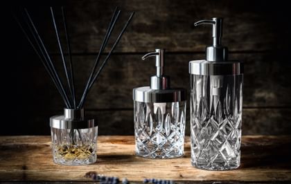 The NACHTMANN Spa Noblesse series on a wooden sideboard. The scent diffuser is filled with a fragrance and eight black sticks. To the right are the small and the tall soap dispenser.<br/>
