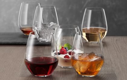 Six SPIEGELAU Authentis Casual tumblers on a wooden table, each of them filled differently. With red wine, with a soft drink on ice, with a creamy desssert, with white wine, with water and ice cubes and with a brown liquor.<br/>