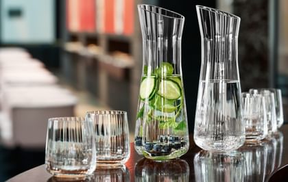 A bar counter with two SPIEGELAU Lifestyle carafes. One of them filled with infused water with blueberries, cucumber and a mint sprig, and one of the pure water. In front and behind the carafes are empty Lifestyle tumblers.<br/>