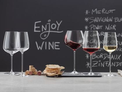 SPIEGELAU Willsberger Anniversary glasses on a marble sideboard. On the left an empty Bordeaux glass and white wine glass, in the middle a pile of corks and potato chips, on the right the filled Burgundy glass, filled red wine glass and filled white wine glass. In the background a blackboard with a menu and the words drink wine.<br/>