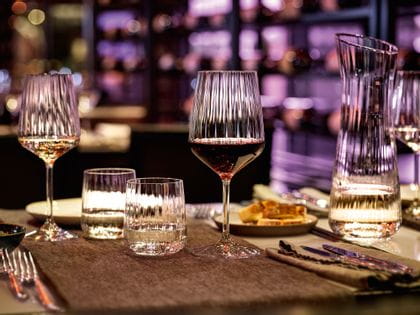 The SPIEGELAU Lifestyle series on a restaurant table in a cosy evening atmosphere. Purple light shines through the smooth wave structure of the filled white wine glass, red wine glass, water tumblers and carafe.<br/>