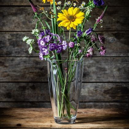 El jarrón de cristal NACHTMANN Carre, lleno de flores moradas, blancas y amarillas sobre un aparador de madera.<br/>