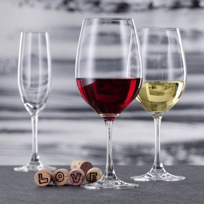 A with red wine filled Bordeaux glass and a with white wine filled white wine glass of the SPIEGELAU Winelovers collection. In the background, there is an empty Champagne flute. Five corks lie behind the Bordeaux glass, four of them showing letters of the word LOVE.<br/>