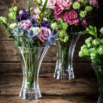 Les vases en cristal NACHTMANN Saphir, remplis de bouquets de fleurs colorés en rouge, vert, violet et bleu sur un buffet en bois.<br/>