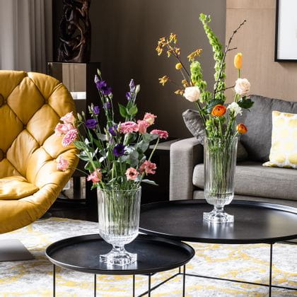 Impresionante, con ramos de flores llenos, jarrones NACHTMANN Minverva sobre mesas auxiliares redondas en un salón. En la sala hay un sofá, una lámpara, un sillón de cuero y una alfombra con estampado amarillo.<br/>