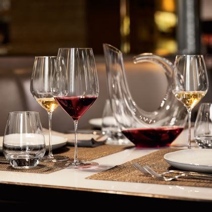 Une table dressée avec des verres SPIEGELAU Highline fabriqués à la main. Le verre à vin rouge est rempli de vin rouge et le verre à vin blanc est rempli de vin blanc. Le gobelet Authentis Casual de SPIEGELAU qui se trouve à côté est rempli d'eau. À l'arrière-plan, on voit une carafe Highline faite à la main et d'autres verres remplis.<br/>