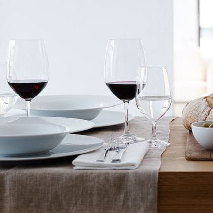 Le verre SPIEGELAU Vino Grande Bordeaux rempli et le verre d'eau minérale sont posés sur une table. Sur les assiettes, il y a des assiettes creuses assorties et, à côté, des couverts et du pain.<br/>