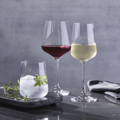 I bicchieri SPIEGELAU Capri su un tavolo di marmo: il bicchiere Bordeaux è riempito di vino rosso, il bicchiere White Wine è riempito di vino bianco e il tumbler è riempito di una bevanda chiara con ghiaccio e un rametto di timo.<br/>
