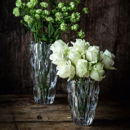 El pequeño jarrón ovalado NACHTMANN Quartz lleno de rosas blancas delante del jarrón alto NACHTMANN Quartz lleno de flores blancas sobre un aparador de madera oscura.<br/>