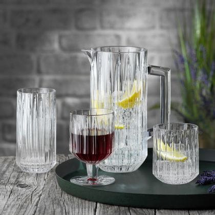 La serie de vasos de cristal NACHTMANN Jules sobre una bandeja redonda para servir. La jarra y el vaso están llenos de agua, cubitos de hielo y rodajas de limón, a su derecha hay lavanda. La copa está llena de vino tinto. El vaso longdrink del fondo está vacío.<br/>