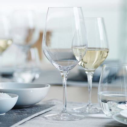 Un verre SPIEGELAU Authentis Bordeaux sur une table posée. Derrière lui, un verre à vin blanc rempli de la ligne Authentis et un gobelet SPIEGELAU Authentis Casual rempli d'eau sont à moitié représentés à droite.<br/>