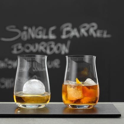 Deux verres à bourbon sans pied SPIEGELAU, remplis de whisky et de glaçons. En arrière-plan, un tableau noir sur lequel est inscrit Single Barrel Bourbon.<br/>