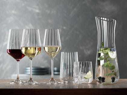 La serie SPIEGELAU Lifestyle su un tavolo. Il bicchiere da vino rosso pieno, il bicchiere da vino bianco pieno, il bicchiere da champagne pieno, un bicchiere da longdrink vuoto, un tumbler pieno d'acqua, cubetti di ghiaccio e una fetta di lime. A destra la caraffa con l'acqua infusa.<br/>