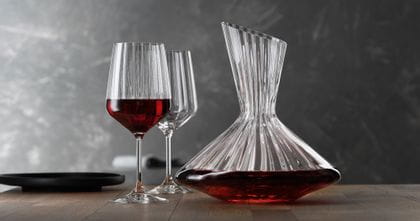 The with red wine filled SPIEGELAU Lifestyle decanter on a table. Left of the decanter two Lifestyle red wine glasses, one of them filled with red wine. On the left edge of the table an empty black plate.<br/>