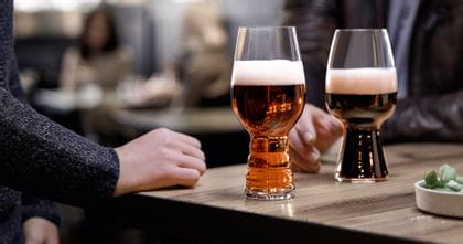 Los vasos de cerveza artesanal SPIEGELAU, el vaso IPA lleno de cerveza IPA y el vaso Stout lleno de cerveza Stout, de pie sobre una mesa de madera. Detrás de ellos, una mano alcanza el vaso de cerveza Stout.<br/>