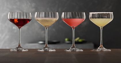 Four SPIEGELAU Lifestyle Coupette glasses on a wooden table. The glasses are filled with different cocktails. From left to right with a Manhattan, a Prosecco, a Cosmopolitan and a Margaritha.<br/>
