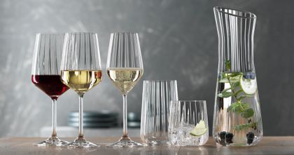 Die SPIEGELAU Lifestyle-Serie auf einem Tisch. Das gefüllte Rotweinglas, das gefüllte Weißweinglas, das gefüllte Champagnerglas, ein leeres Longdrinkglas, ein Becher gefüllt mit Wasser, Eiswürfeln und einer Limettenscheibe. Rechts die Karaffe mit gefülltem Wasser.<br/>