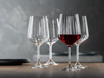 Four SPIEGELAU Lifestyle red wine glasses on a wooden table. One of them is filled with red wine.<br/>