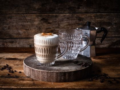 Due tazze per bevande calde NACHTMANN Ethno su un tagliere in legno. Una tazza è piena di cappuccino, quella dietro è vuota. Sullo sfondo, una macchina da caffè italiana.<br/>