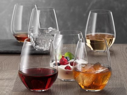 Six SPIEGELAU Authentis Casual tumblers on a wooden table, each of them filled differently. With red wine, with a soft drink on ice, with a creamy desssert, with white wine, with water and ice cubes and with a brown liquor.<br/>