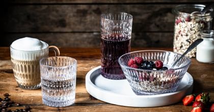 The NACHTMANN Ethno series on a wooden table. The hot beverage mug is filled with cappuccino, the tumbler with mineral water. The longdrink glass, filled with berry juice and the bowl, filled with a crunchy muesly, are standing on a white serving tray. In the background a jar with cereals and milk.<br/>