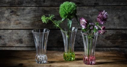 Small NACHTMANN vases SPRING on a wooden table. One has a lime colored bottom and green flowers in it, the other one has a rosé colored bottom and purple flowers in it.<br/>