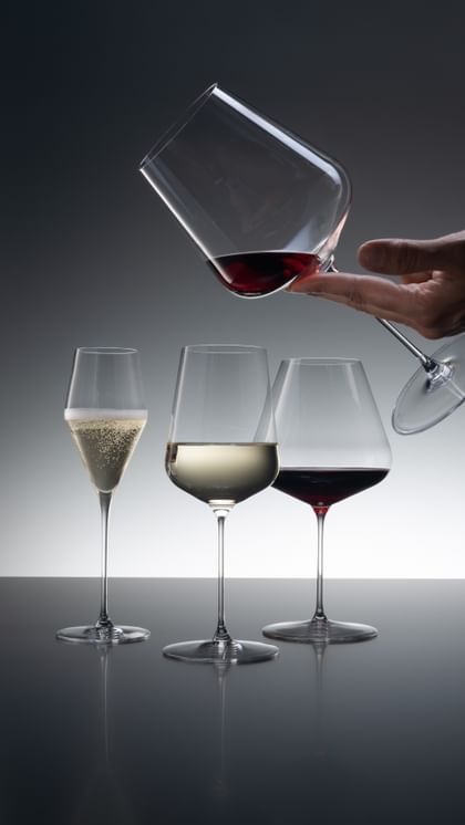 Drei SPIEGELAU Definition Gläser, von denen das Champagnerglas mit Sekt, das Universalglas mit Weißwein und das Burgunderglas mit Rotwein gefüllt ist. Von der rechten oberen Ecke kommend hält eine Hand das Bordeaux-Glas, das mit etwas Rotwein gefüllt ist.<br/>