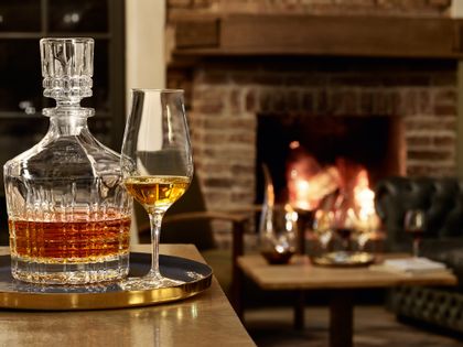 A cozy fireplace with a with Whisky filled SPIEGELAU Perfect Serve Collection Whisky Carafe and a filled SPIEGELAU Whisky Snifter Premium on a serving tray in the foreground. <br/>