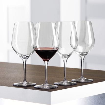 Vier SPIEGELAU Authentis Bordeaux-Gläser auf einem Tisch, eines davon ist mit Bordeaux-Wein gefüllt.<br/>