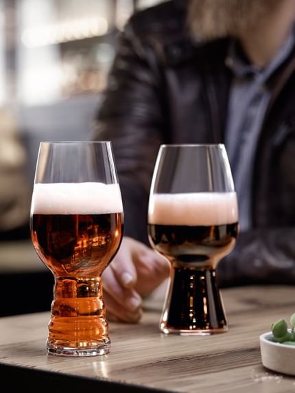 The SPIEGELAU Craft Beer Glasses IPA glass filled with IPA beer and the Stout glass filled with Stout beer standing on a wooden table. Behind them a hand reaching for the Stout glass.<br/>