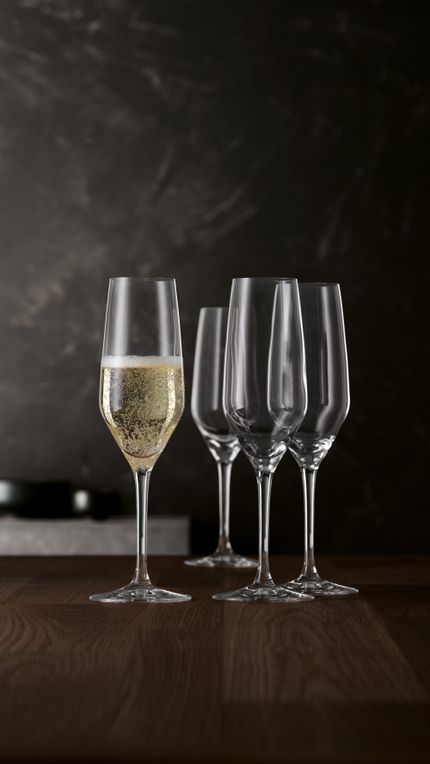 Four SPIEGELAU Style Champagne flutes on a wooden table. One glass is filled with sparkling wine.<br/>